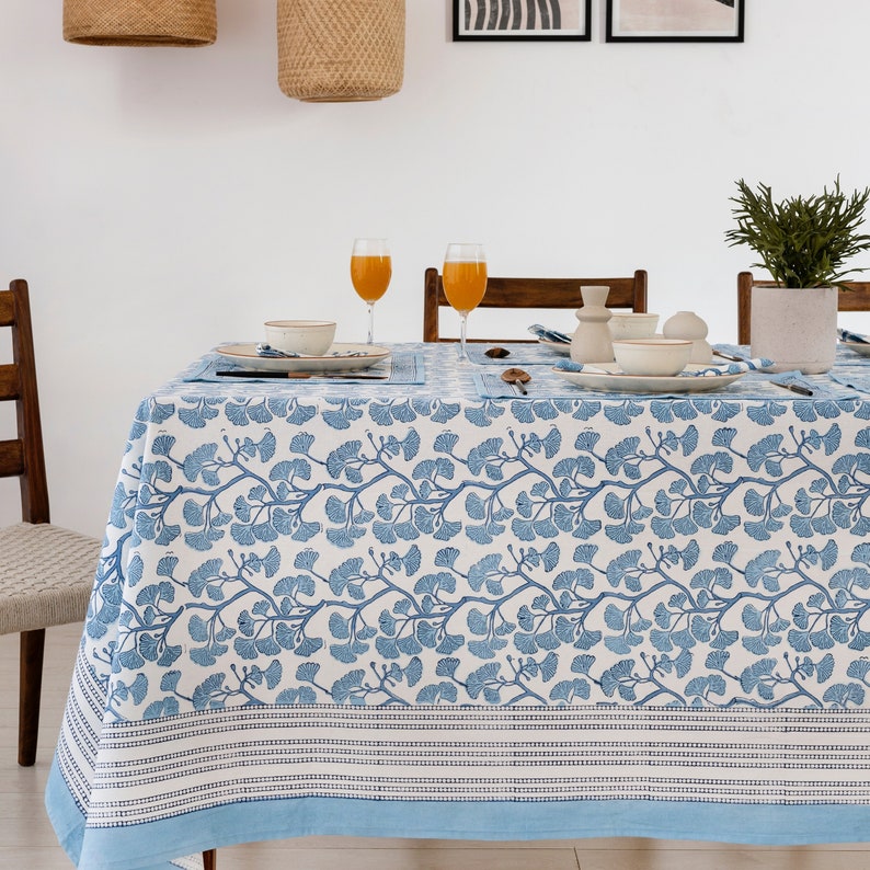 Puede incluir: Un mantel blanco con un patr&oacute;n de hojas de ginkgo azul. El mantel tiene un borde azul con rayas blancas. El mantel est&aacute; puesto para una comida con dos vasos de zumo de naranja, platos y cubiertos.