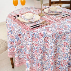 May include: A round white tablecloth with a pink and blue floral pattern. The tablecloth has a red border with a geometric design. Two place settings are set on the tablecloth with white plates, bowls, and napkins.