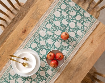 May include: Overhead shot of a wooden table set for a meal. A green floral patterned table runner is centered on the table. White plates, gold silverware, and a bowl of pomegranates are arranged on the table. Two pomegranates are scattered on the runner.