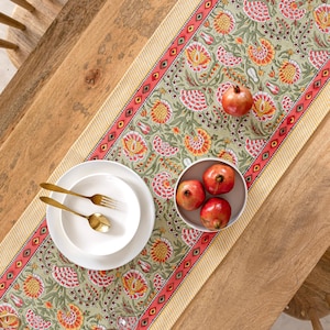 Pode incluir: Um caminho de mesa floral com fundo verde claro e um padrão repetido de flores vermelhas e amarelas. O caminho de mesa está sobre uma mesa de madeira, com pratos brancos, talheres dourados e uma tigela de romãs.