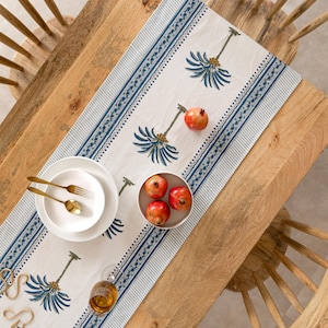 May include: A table setting featuring a cream-colored table runner with a blue floral pattern and striped borders. The table is set with white plates, gold silverware, and a bowl of red pomegranates. A glass of amber liquid sits on the table.