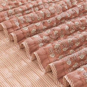 May include: A quilted cotton blanket in a dusty rose color featuring a repeating white floral block print pattern. The blanket has a finished edge with a white and dusty rose diagonal striped border and a matching striped underside.