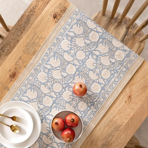 May include: A blue floral table runner with a cream-colored flower pattern, placed on a wooden table. The runner is decorated with a cream-colored border. Two white plates with gold utensils and a bowl of pomegranates are on the table.