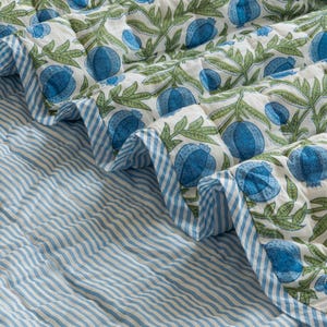 Op de afbeelding: Een gewatteerde sprei met een witte achtergrond, met een patroon van blauwe granaatappels en groene bladeren. De rand heeft een blauw-wit gestreept ontwerp. Het onderste gedeelte heeft een blauw-wit gestreept patroon.