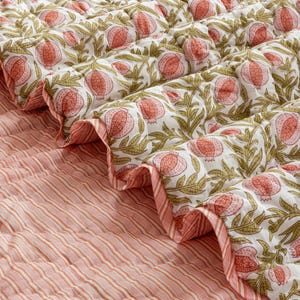 Może przedstawiać: Pikowana narzuta na łóżko z wzorem granatu na białym tle i oliwkowymi liśćmi. Druga strona ma różowo-biały wzór w paski. Krawędzie są z falbankami z różowo-białym paskiem.