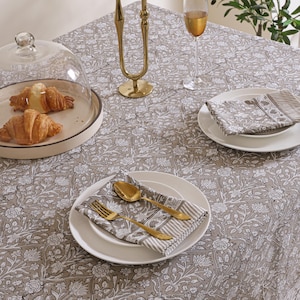 May include: A taupe and white floral patterned tablecloth featuring a repeating botanical design. The table is set with white plates, folded napkins, gold-toned cutlery, a glass cloche containing two croissants, and a tall gold-toned candle holder.