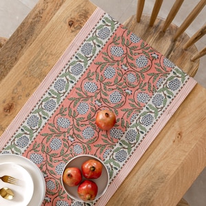 Op de afbeelding: Een koraalkleurige tafelloper met een bloemenpatroon met blauwe en groene accenten. De loper is versierd met een herhalend ontwerp van gestileerde bloemen en bladeren. De loper ligt op een houten tafel, met granaatappels en een kom met granaatappels.