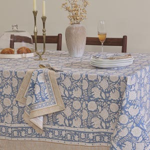 Puede incluir: Un mantel rectangular con un estampado floral azul y blanco con motivos de elefantes y un borde a rayas. Una servilleta a juego descansa sobre la superficie, decorada con un jarrón, velas y una pila de platos blancos.