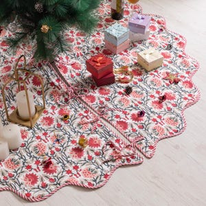 May include: A white Christmas tree skirt featuring a red floral design and a scalloped border. The skirt is adorned with small gift boxes, decorations, and a gold candle holder. Designed to encircle the base of a Christmas tree.