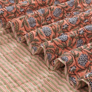 May include: A quilted textile featuring a peach-colored top layer with a repeating dark blue and green floral pattern. The underside of the fabric displays a light green and white striped pattern, visible where the quilt is folded over.