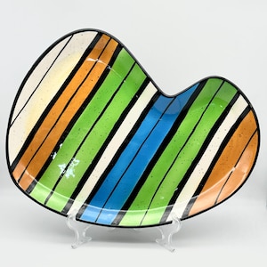 May include: A colorful, heart-shaped ceramic serving dish with a black rim. The dish features diagonal stripes in yellow, orange, green, white, and blue, separated by black lines. The dish is displayed on a clear stand.