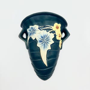 May include: A dark teal wall pocket vase with a floral design. The vase has a curved shape with two handles and a textured surface. The floral design features white and blue leaves with yellow flowers.