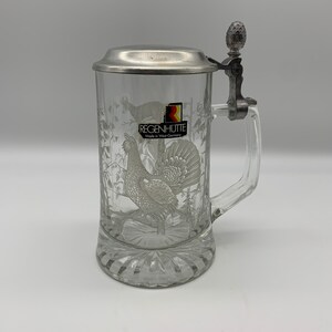 May include: A clear glass beer stein with a pewter lid and handle. The stein features an etched design of a grouse in a forest setting. The lid has a small pewter finial in the shape of a pine cone. The stein is labeled "Regenh&uuml;tte Made in West Germany".