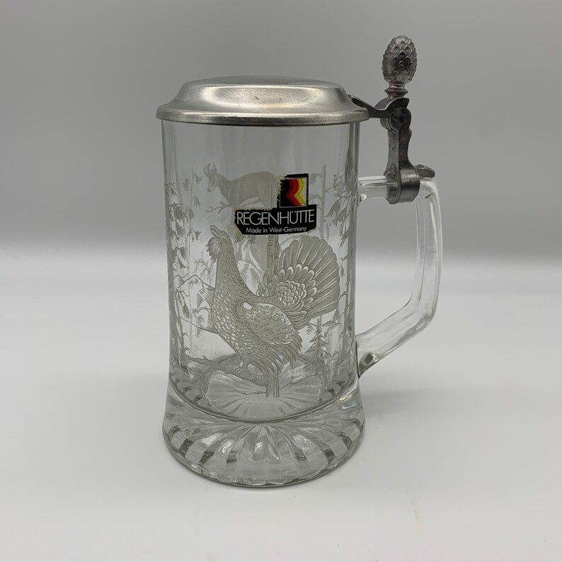 May include: A clear glass beer stein with a pewter lid and handle. The stein features an etched design of a grouse in a forest setting. The lid has a small pewter finial in the shape of a pine cone. The stein is labeled "Regenh&uuml;tte Made in West Germany".