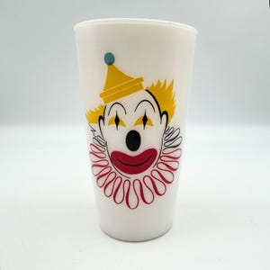 May include: A white tapered drinking glass featuring a graphic illustration of a clown face. The clown has a yellow hat with a blue dot, yellow hair, black diamond-shaped eyes, a black nose, and a red mouth surrounded by a red ruffled collar.