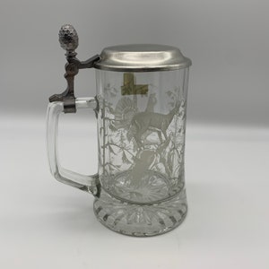May include: A clear glass beer stein with a pewter lid and a handle. The stein is etched with a white design of a deer and a turkey in a forest setting.