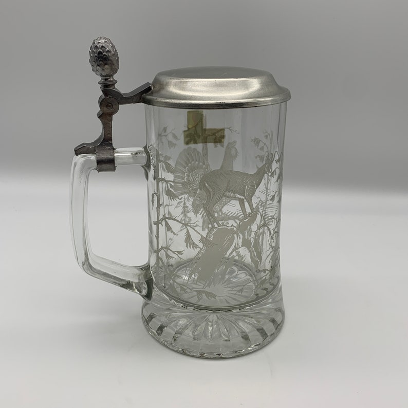 May include: A clear glass beer stein with a pewter lid and a handle. The stein is etched with a white design of a deer and a turkey in a forest setting.