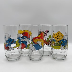 May include: Set of four Smurfs themed drinking glasses. Each glass features a different Smurf character: Brainy, Papa, Lazy, and Grouchy. The glasses are clear with colorful cartoon illustrations.