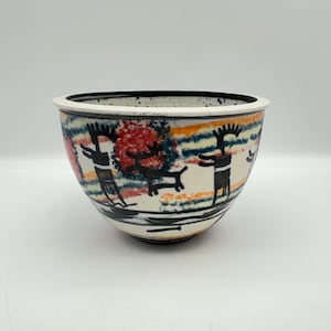 May include: A ceramic bowl with a wide rim and a rounded body. The bowl features a hand-painted design with black figures, red and orange trees, and colorful horizontal stripes. The rim is white with black speckles.