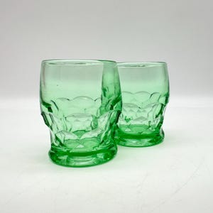 May include: Two green-tinted drinking glasses. The glasses have a short, rounded base and a textured, diamond-like pattern on the lower half. The rims have a scalloped design. The glasses are empty and sit on a white surface.