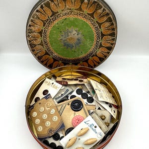 May include: A round metal tin with a decorative lid featuring orange tulip motifs and a green center, filled with various sewing buttons on cardboard display cards. The cards include brands like Le Chic and Le Bouton with assorted button shapes.