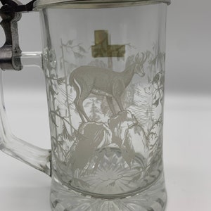 May include: A clear glass beer stein with a pewter lid and a white etched design of a deer in a forest setting.