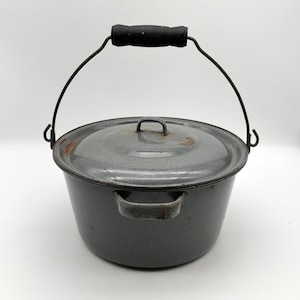 May include: A gray enamelware pot with a lid and a wire handle with a black wooden grip. The pot has two side handles and a small handle on the lid. The pot is likely vintage or antique.