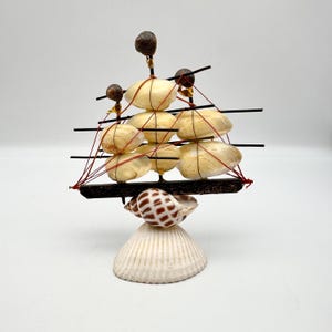 May include: A decorative seashell ship sculpture. The ship features a base made of a large, white seashell, topped with a smaller shell. The ship's sails are made of cream-colored shells, and the rigging is red. The masts are black.