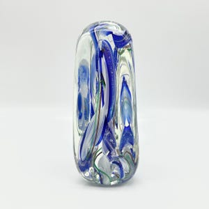 May include: A clear glass art piece with blue, white, and green swirls. The elongated, rounded shape features a mix of colors and textures, creating a unique decorative item. The glass is transparent, allowing light to pass through.