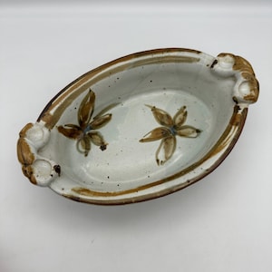 May include: Oval ceramic serving dish with a speckled white interior and brown floral design. The rim and handles are brown. The dish measures approximately 10 inches long and 6 inches wide.