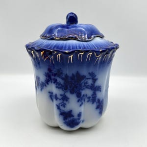 May include: A white porcelain sugar bowl with a scalloped rim and a matching lid. The exterior features a deep blue floral transfer pattern that fades into the white base. The rim and lid edges are accented with delicate gold painted trim.