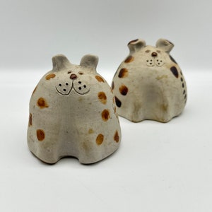 May include: Two ceramic dog figurines. The dogs are off-white with brown spots and simple facial features. They have small ears and are sitting upright. The figurines have a rustic, handmade appearance and are set against a white background.