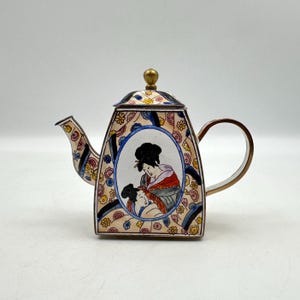 May include: A metal teapot featuring a central oval medallion with an illustration of two figures. The body is decorated with a colorful floral pattern on a cream background, accented by dark blue stripes, a curved handle, and a gold-toned lid knob.