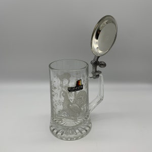 May include: A clear glass beer stein with a pewter lid and handle. The stein features an etched design of a rooster and a deer. The lid is open and the stein is sitting on a white surface. The stein has the text "Regenh&uuml;tte Made in West Germany" on the front.