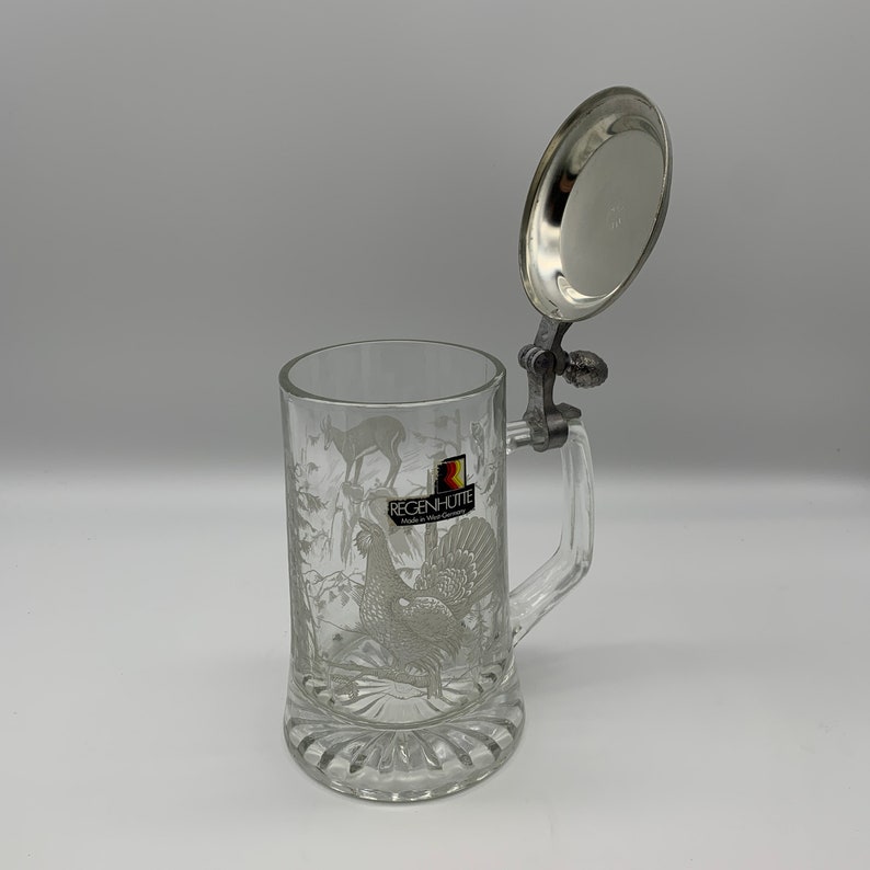 May include: A clear glass beer stein with a pewter lid and handle. The stein features an etched design of a rooster and a deer. The lid is open and the stein is sitting on a white surface. The stein has the text "Regenh&uuml;tte Made in West Germany" on the front.