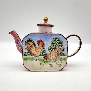 May include: A decorative teapot with a pink lid and spout, featuring a hand-painted scene of a rooster, hen, and chicks in a field. The teapot has a gold handle and a square body with a colorful design.