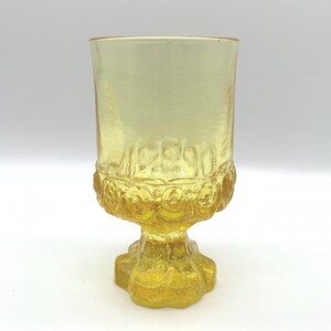 May include: A vintage yellow glass goblet with a textured, patterned design. The goblet has a wide bowl, a decorative base, and a stem. The glass has a slight amber hue, and the base is textured. The goblet is likely used for serving beverages.