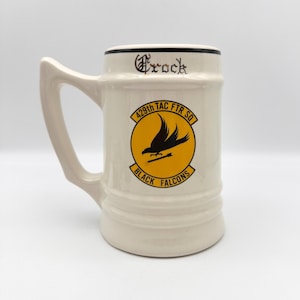 May include: Cream-colored ceramic beer stein with a large handle. The stein features the text "Brock" in black script and a circular emblem with a black bird on a yellow background. The emblem reads "429th TAC FTR SQ" and "BLACK FALCONS".