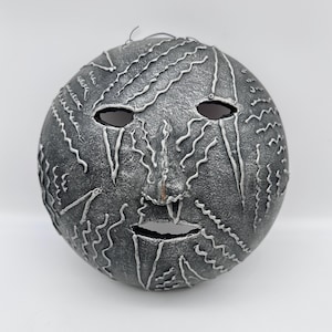 May include: A gray, spherical mask with a textured surface and white, wavy accents. The mask has eye and mouth openings, and a nose. The design resembles a face with a serene expression. The mask is likely for decorative or theatrical purposes.