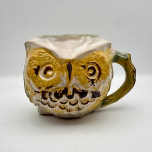 May include: A ceramic mug shaped like an owl with a textured surface. The owl features large yellow eyes with dark brown pupils, a dark brown beak, and brown feather details on a cream-colored body. A curved handle extends from the right side.
