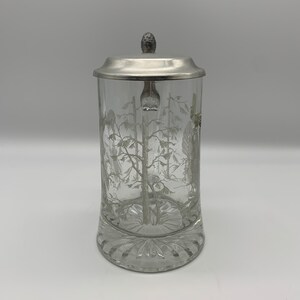 May include: A clear glass beer stein with a pewter lid and a white etched design of trees. The stein has a textured base.