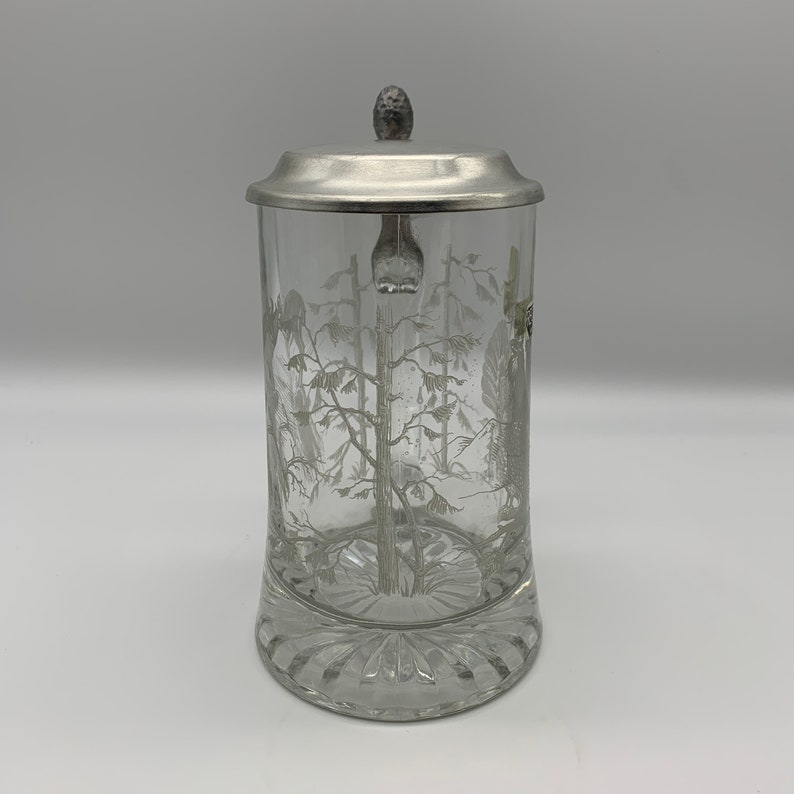 May include: A clear glass beer stein with a pewter lid and a white etched design of trees. The stein has a textured base.