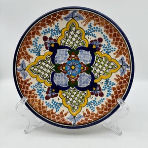 May include: A round ceramic plate featuring a central blue flower surrounded by intricate geometric patterns in yellow, dark blue, green, and brown. The border displays a repeating brown scale-like design against a cream-colored background.
