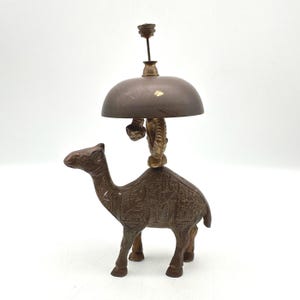 May include: A vintage brass camel figurine with an attached bell. The camel is brown with etched details, supporting a bell with a dark brown dome. The bell is attached to a decorative brass piece. The background is white.