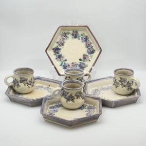 May include: A ceramic tea set featuring a cream-colored hexagonal serving tray and matching small plates and mugs. The set is decorated with a floral pattern in shades of purple and blue, with a light purple border.