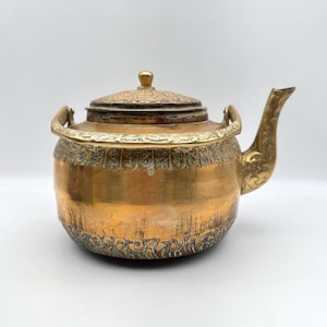 May include: A vintage brass teapot with intricate floral patterns. The teapot features a curved spout, a handle on each side, and a decorative lid with a small knob. The body of the teapot has a rounded shape and is made of brass.