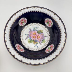May include: A dark blue ceramic serving bowl with a white scalloped edge and gold trim. The bowl features a central floral design and smaller floral accents. The flowers are pink, purple, and yellow.