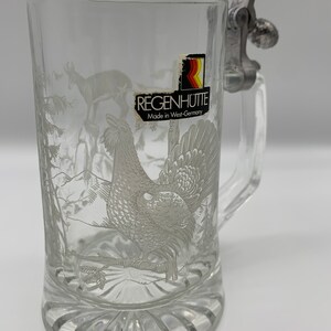 May include: A clear glass beer stein with a pewter lid and a white etched design of a grouse in a snowy forest. The stein has a label that reads "Regenh&uuml;tte Made in West-Germany".
