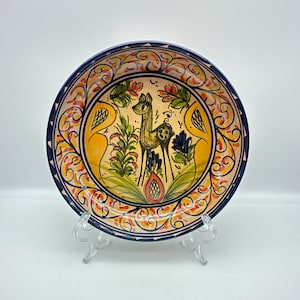 May include: A ceramic plate with a blue and white border and a colorful floral design featuring a deer in the center. The plate is decorated with yellow, red, green, and blue colors.