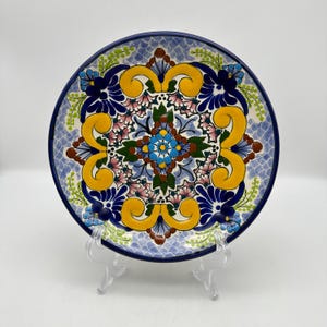 May include: A round ceramic plate featuring a vibrant hand-painted design with a central blue floral motif, surrounded by yellow scrolls, green leaves, and pink accents. The background has a light blue scale pattern with a dark blue rim border.